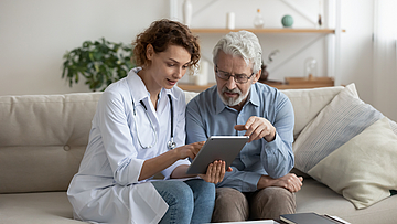 Eine Ärztin und ihr alterer Patient sitzen auf einem Sofa. Die Arztin hält ein Tablett. Sie erklärt dem Patienten die Ergebnisse einer Untersuchtung.