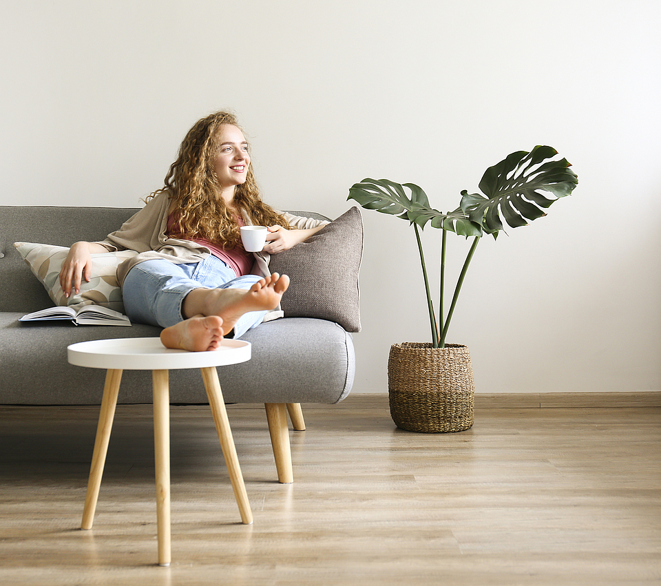 Lächende Frau sitzt auf dem Sofa und hat die Beine auf den Tisch gelegt. Sie hält eine Kaffetasse in der linken Hand.