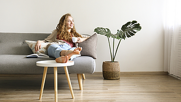 Lächende Frau sitzt auf dem Sofa und hat die Beine auf den Tisch gelegt. Sie hält eine Kaffetasse in der linken Hand.