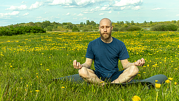 Ein Mann sitzt auf einer Blumenwiese er meditiert im Schneidersitz auf der Yogamatte.