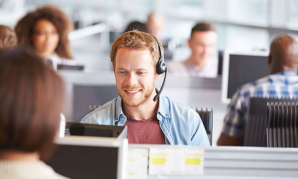 In einem Callcenter sitzt ein junger, blonder Mann mit Jeanshemd. Er hat ein Headset auf und telefoniert.