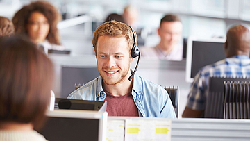 In einem Callcenter sitzt ein junger, blonder Mann mit Jeanshemd. Er hat ein Headset auf und telefoniert.