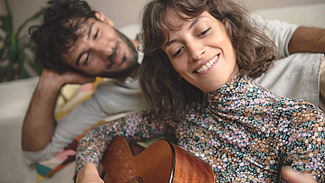 Junges Paar glückliches liegt auf einem Bett, sie spielt Gitarre und er schaut ihr dabei zu.