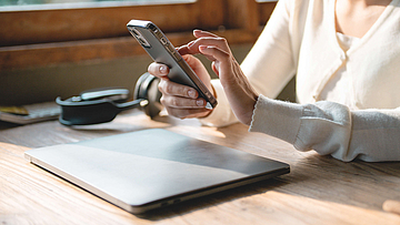 Frau tippt in das Smartphone, vor ihr auf dem Tisch liegt ein Laptop.