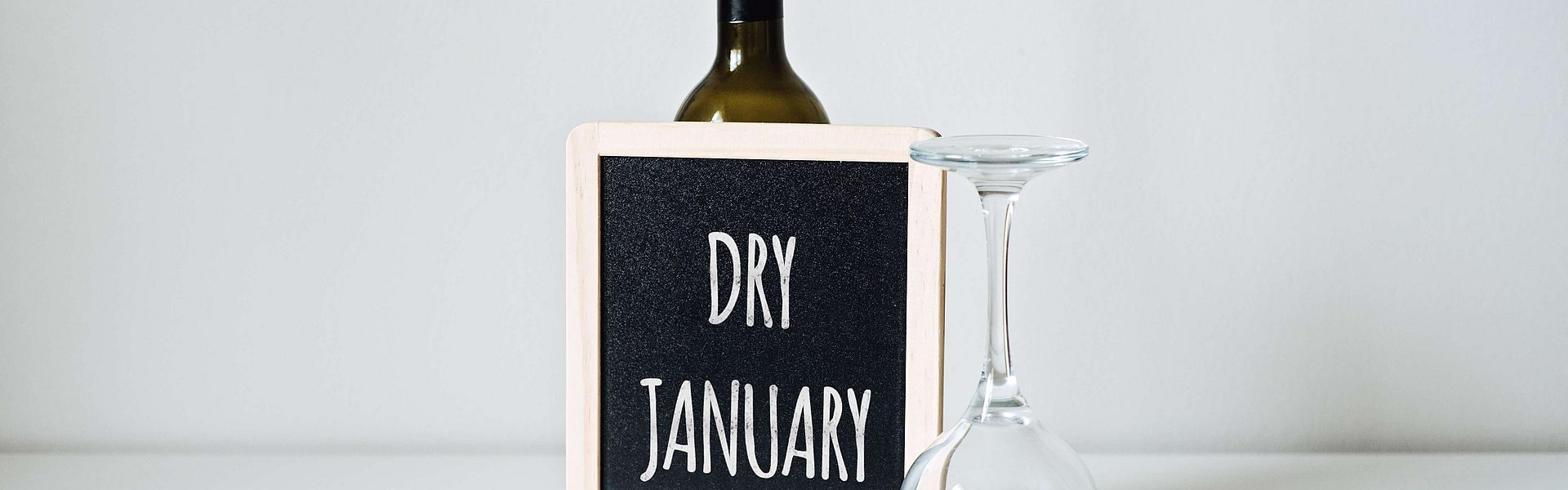 Schild mit Aufschrift „Dry January” lehnt an geschlossener Flasche, neben einem umgedrehten Weinglas stehend