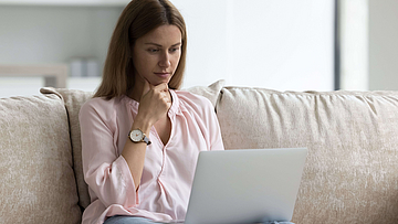 Eine Frau sitzt auf einem Sofa. Auf ihrem Schoß liegt ein aufgeklappter Laptop.