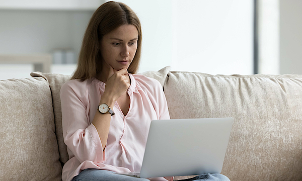 Eine Frau sitzt auf einem Sofa. Auf ihrem Schoß liegt ein aufgeklappter Laptop.