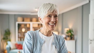 Reife freundlich lächelnde Frau steht mit gekreuzten Armen im Raum.