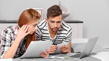 Ein Mann und eine Frau liegen auf dem Bett. Vor ihnen ist ein Laptop. Sie halten Unterlagen in Händen. Sie machen ihre Steuererklärung.