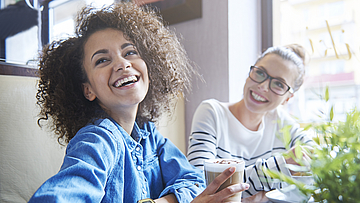 Zwei glückliche Frauen sind in einem Bistro. Die Frau im Vordergrund trinkt einen Latte Macchiato.