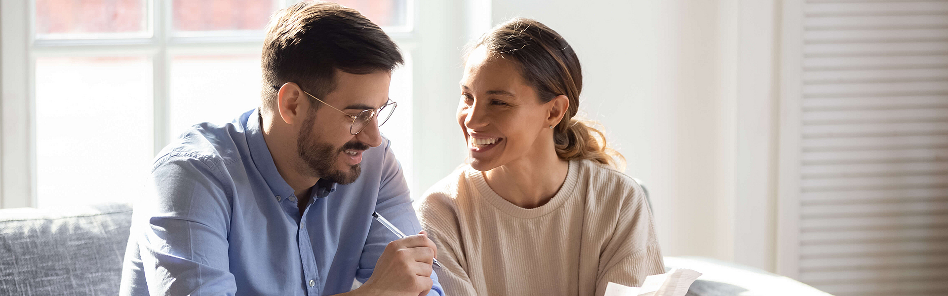 Paar sitzt auf dem Sofa auf dem Tisch steht ein Laptop. Der Mann spricht und die Frau lacht.