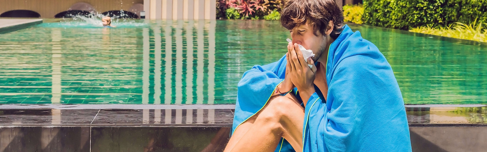 Kranker Reisender. Der Mann hat sich im Urlaub erkältet, sitzt traurig am Pool, trinkt Tee und putzt sich die Nase in eine Serviette.