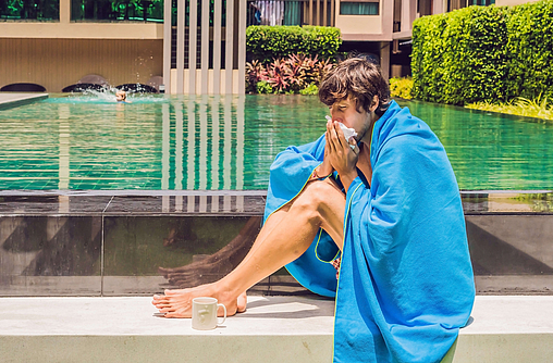 Kranker Reisender. Der Mann hat sich im Urlaub erkältet, sitzt traurig am Pool, trinkt Tee und putzt sich die Nase in eine Serviette.