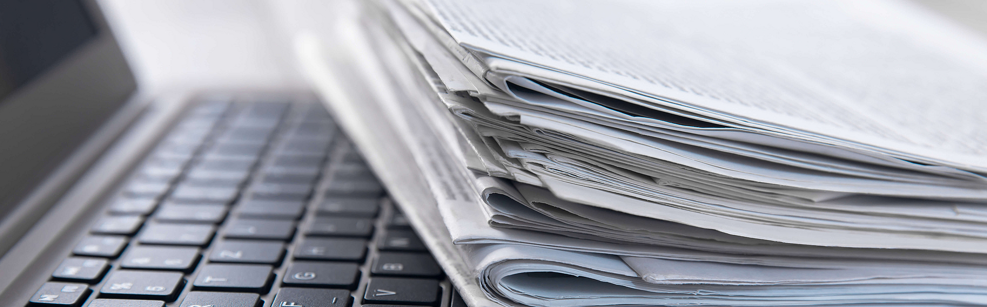 Headerfoto_Presse-Übersichtsseite_Zeitungen_Laptop Stapel Zeitungen liegt auf der Tastatur eines geöffneten Laptops
