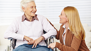 Eine ältere Dame sitzt lächelnd im Rollstuhl, sie unterhält sich mit einer jungen Frau.