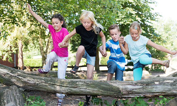 Kinder im Wald hüpfen über einen Baumstamm.