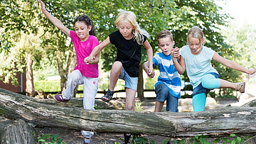 Kinder im Wald hüpfen über einen Baumstamm.