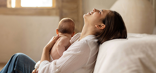 Frau mit Wochenbettdepression sitzt auf dem Boden und versucht zu entspannen. Sie hat dabei ein Baby im Arm.