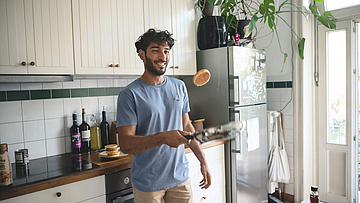 Mann steht in der Küche. Er wendet einen Pfannkuchen in der Pfanne.