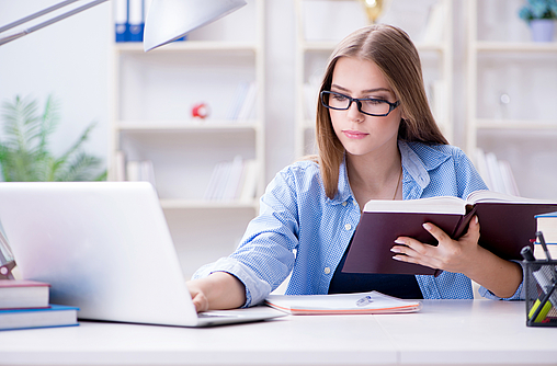 Eine junge Frau sitzt vor einem Laptop. Sie hat ein Buch in der Hand und macht sich Notizen. Sie lernt.