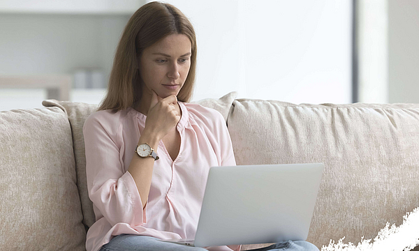 Junge Frau schaut nachdenklich und konzentriert auf ihren Laptop. Sie sitzt auf einem Sofa.