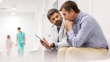 Ein Mann und sein Arzt sitzen im Gang eines Krankenhaus. Er erklärt dem Mann etwas.