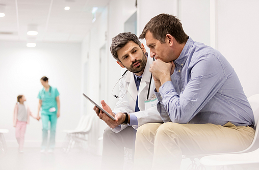 Ein Arzt und ein Mann sitzen nebeneinander auf einem Gang im Krankenhaus. Der Arzt hält ein Tablet in der Hand. Er erklärt dem Mann etwas. Der Mann schaut bestürzt.