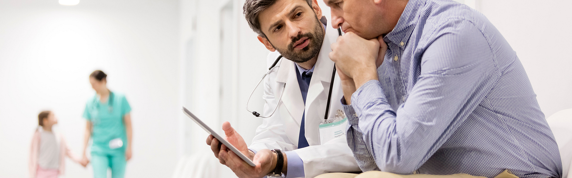 Ein Arzt und ein Mann sitzen nebeneinander auf einem Gang im Krankenhaus. Der Arzt hält ein Tablet in der Hand. Er erklärt dem Mann etwas. Der Mann schaut bestürzt.