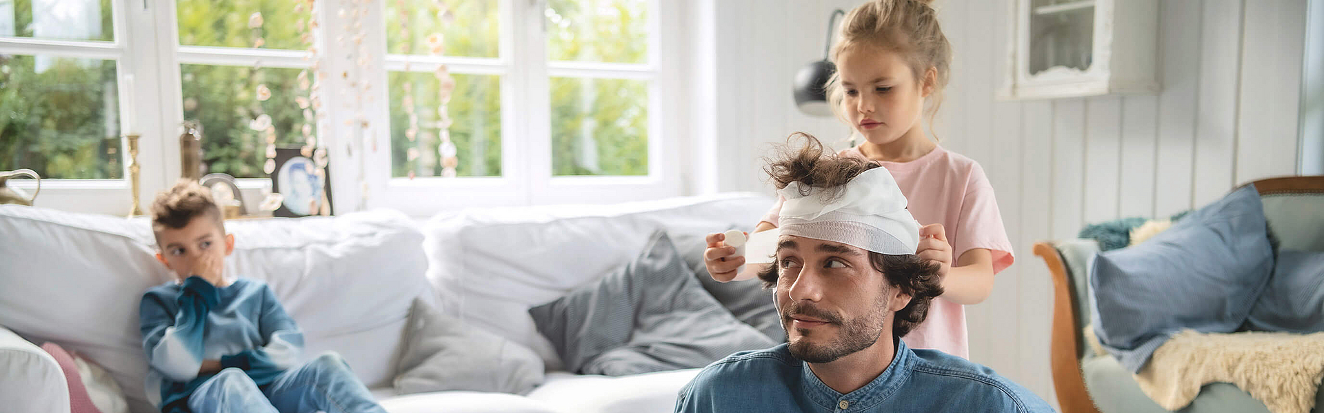 Das Mädchen verbindet mit einer weißen Mullbinde Vaters Kopf. Dabei sitzt er im Wohnzimmer auf dem Fußboden, das Mädchen steht hinter ihm. Auf dem Sofa sitzt ein kleiner Junge.