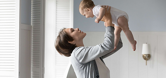 Frau am Fenster hebt ihr Baby in die Höhe