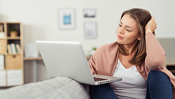 Frau sitzt auf dem Sofa und arbeitet mit dem Laptop. Der Laptop ist auf dem Sofa abgelegt.