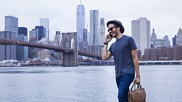 Ein Mann vor der Skyline von New York. Er trägt eine Laptop-Tasche und telefoniert.