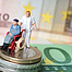 Ein Rollstuhlfahrer mit Pflegepersonal steht auf mehren Geldscheinen