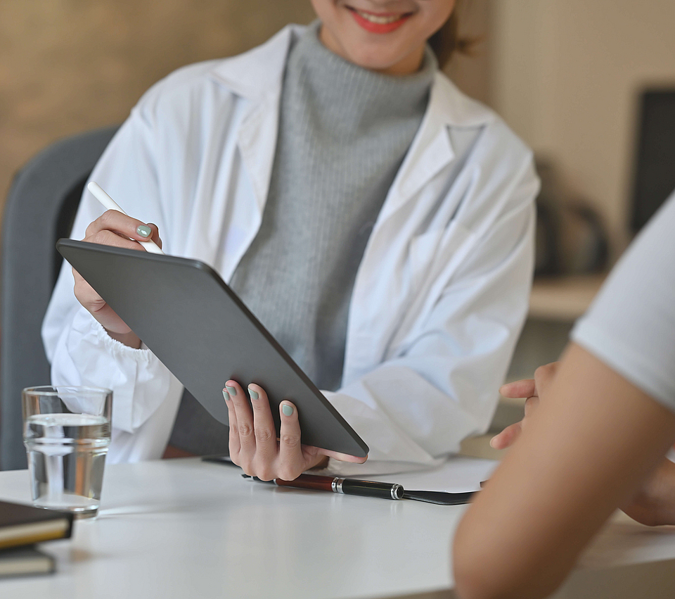 Ein Arzt und sein Patient sitzen an einem Tisch. Der Arzt hält ein Tablet in der Hand und zeigt darauf etwas dem Patienten.