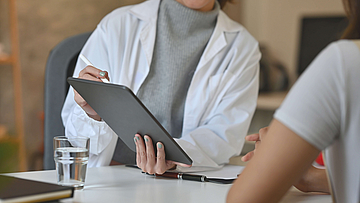 Ein Arzt und sein Patient sitzen an einem Tisch. Der Arzt hält ein Tablet in der Hand und zeigt darauf etwas dem Patienten.