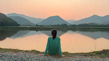 Eine Frau sitzt am Ufer eines Sees. Sie schaut aufs Wasser. Die Sonne geht unter. Sie ist alleine.