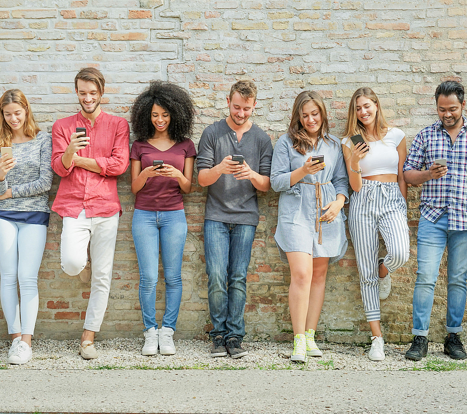 Eine Gruppe junger Menschen steht vor einer Mauer. Jeder hat ein Smartphone in der Hand und blickt darauf.