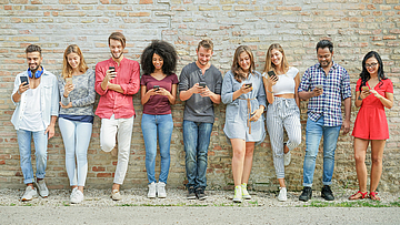 Eine Gruppe junger Menschen steht vor einer Mauer. Jeder hat ein Smartphone in der Hand und blickt darauf.