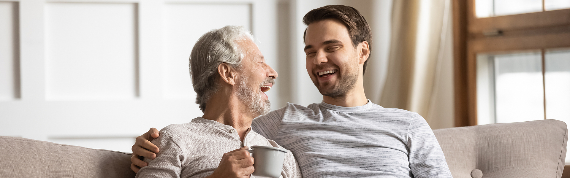 Ein Rentner und ein Mann sitzen lachend auf dem Sofa.